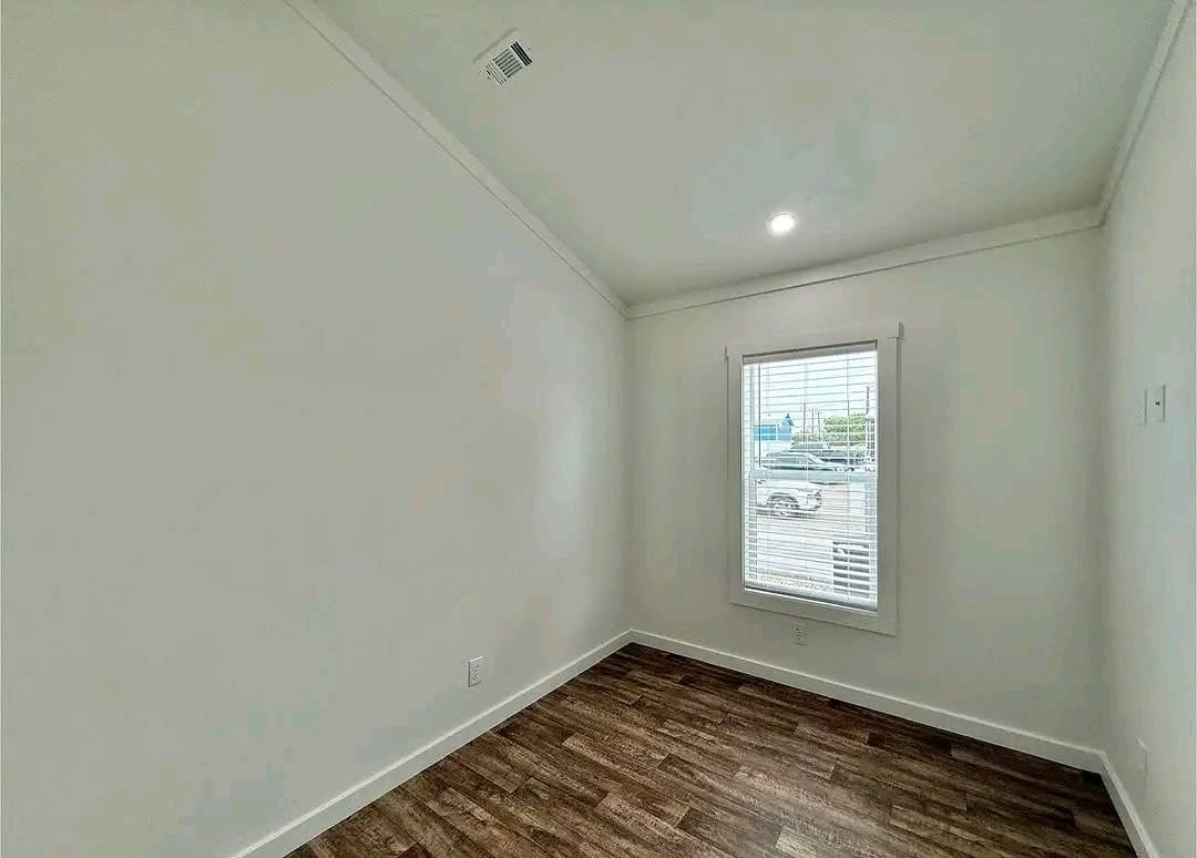 A compact, empty room with white walls and dark wood flooring. It features a window with blinds showing a parking lot view. The room feels simplistic and bright.