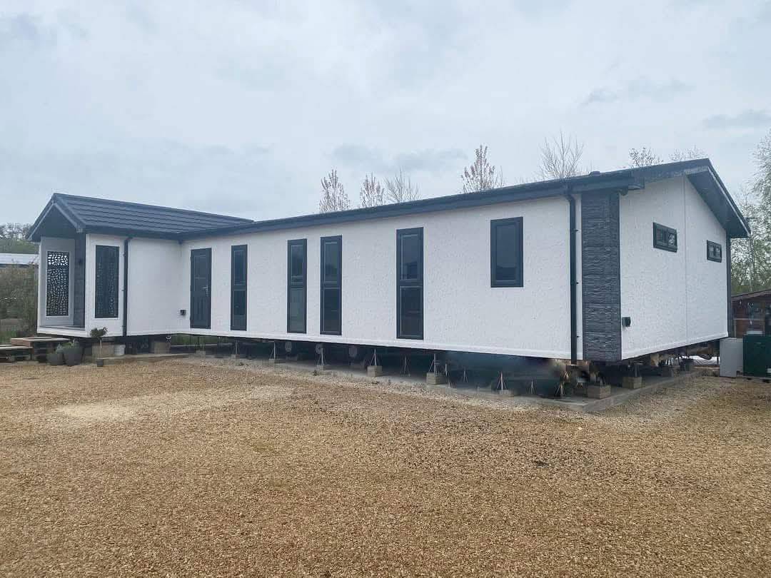 Modern, rectangular white house with black-framed windows on raised supports, set against a cloudy sky, conveying a minimalist, contemporary style.