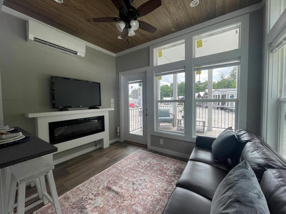 Cozy living room with gray walls, wooden ceiling, and a ceiling fan. Features a leather sofa, wall-mounted TV, electric fireplace, and large windows showing a patio and trees.