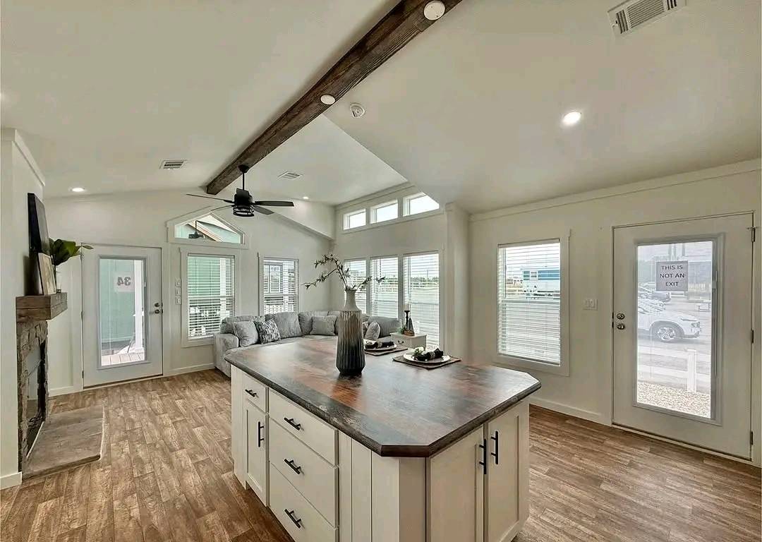 Modern kitchen and living area with wood flooring and ceiling beams. Features a central island with decor, large windows, and a cozy corner sofa. Bright and inviting ambiance.