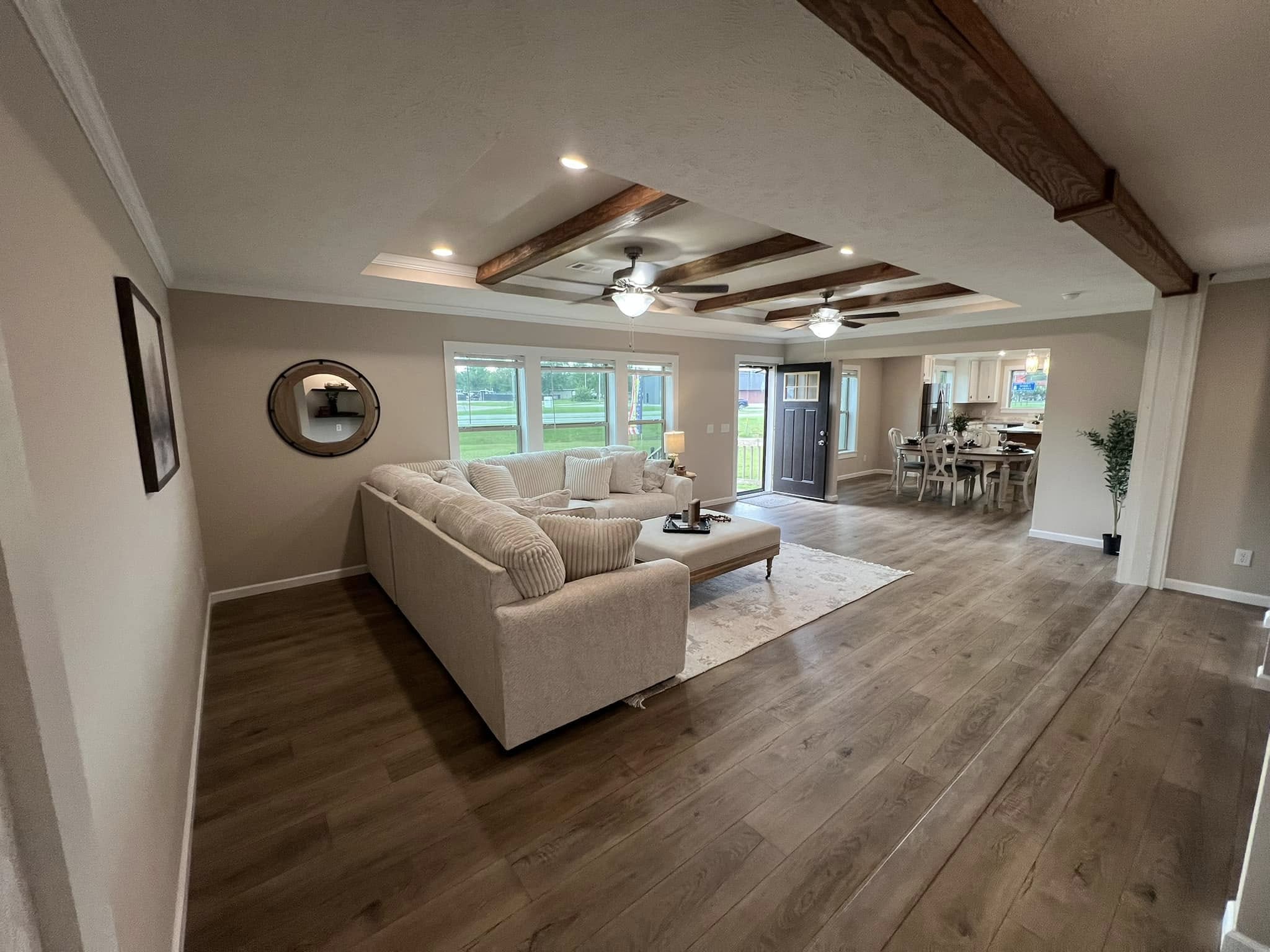 Spacious living room with wooden beams, beige sectional sofa, and rug. Natural light from large windows and open layout leading to dining area. Cozy and welcoming.