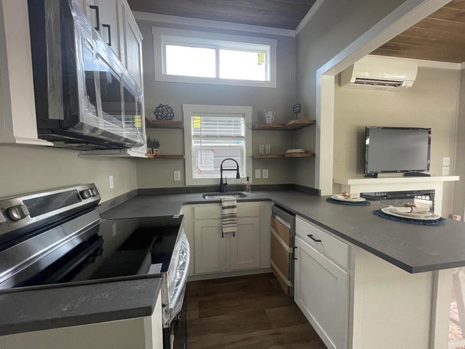 Modern kitchen with gray countertops, white cabinets, and stainless steel appliances. Open shelves display decor. TV area visible in the background.