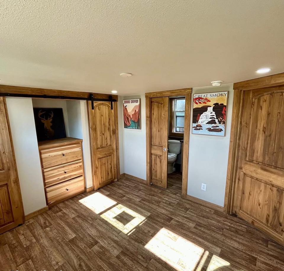 Cozy room with wooden floors and doors, showcasing a dresser and colorful landscape posters. Warm sunlight filters in, creating a rustic, inviting atmosphere.