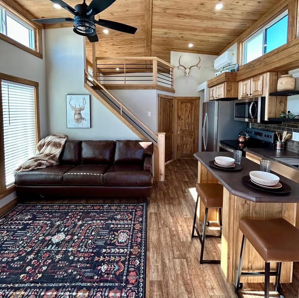 Cozy living room with wood accents, featuring a leather sofa, decorative rug, staircase with railing, and a kitchen island. Warm, inviting atmosphere.