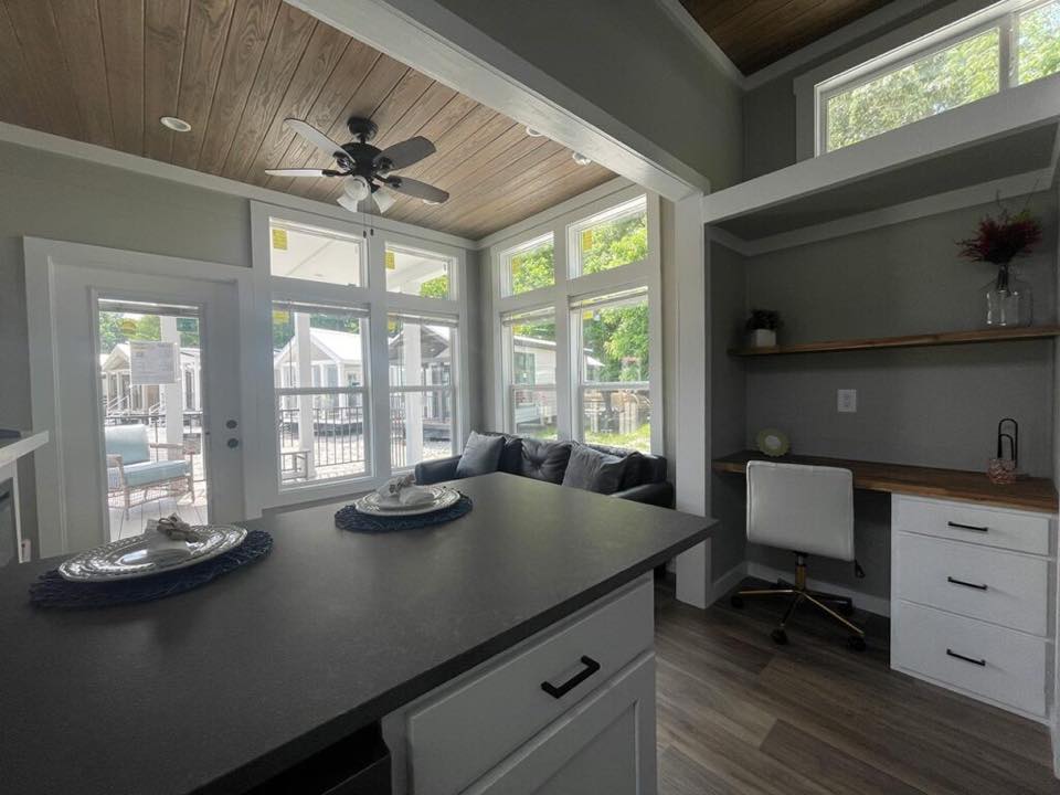 Modern room with large windows, bright light, and a cozy sofa. A kitchen island in the foreground, with an office nook featuring a desk and chair.
