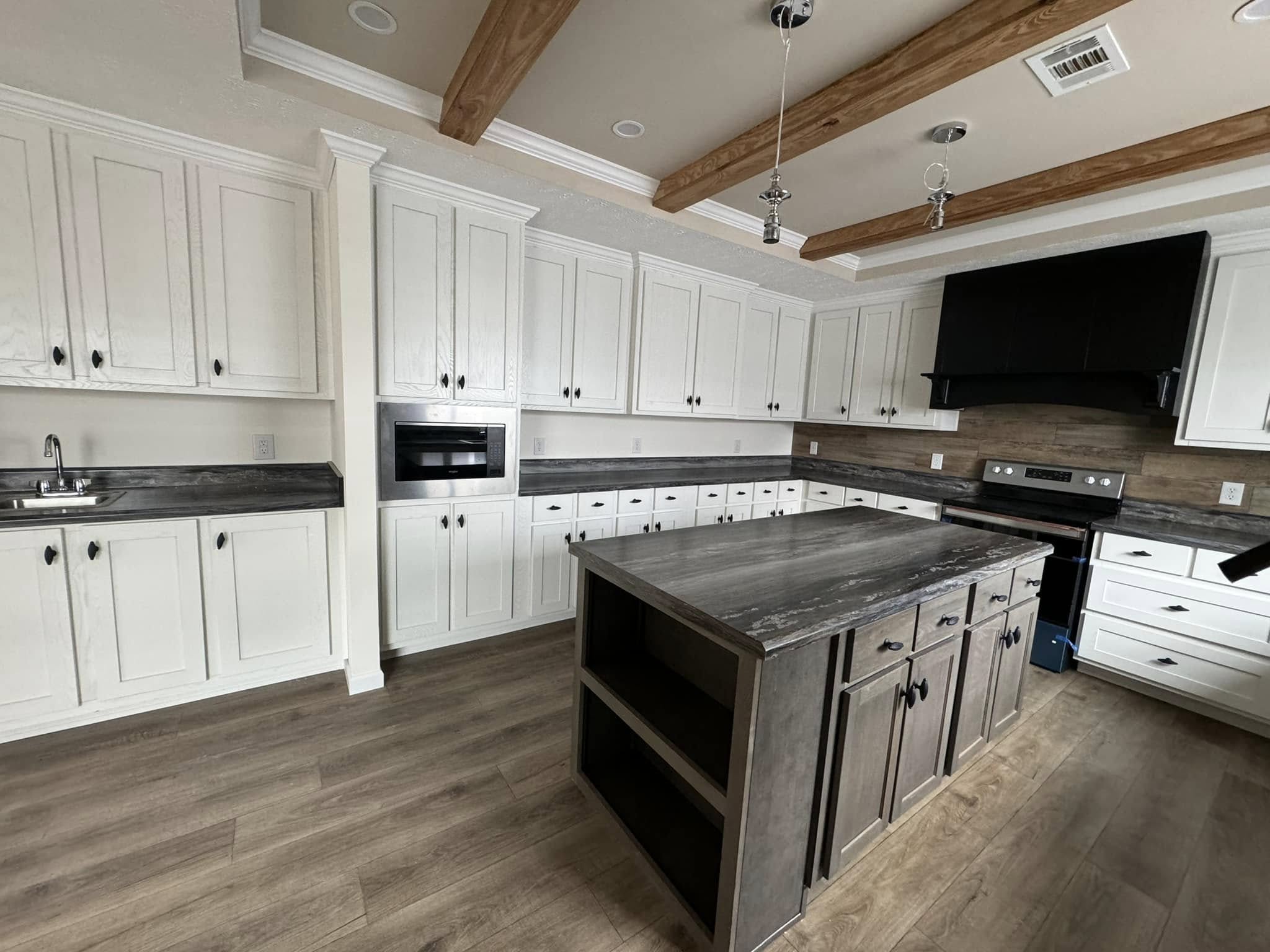 Spacious kitchen with white cabinets, black counters, and a central island. Wooden beams and gray tiles create a modern, rustic feel.
