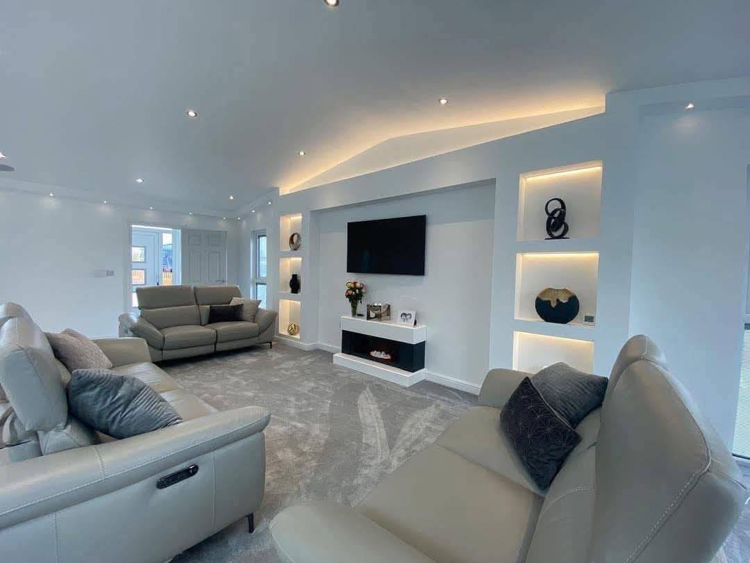 Modern living room with gray leather sofas, a wall-mounted TV, built-in illuminated shelves with decor, and a soft gray carpet. Bright and stylish ambiance.