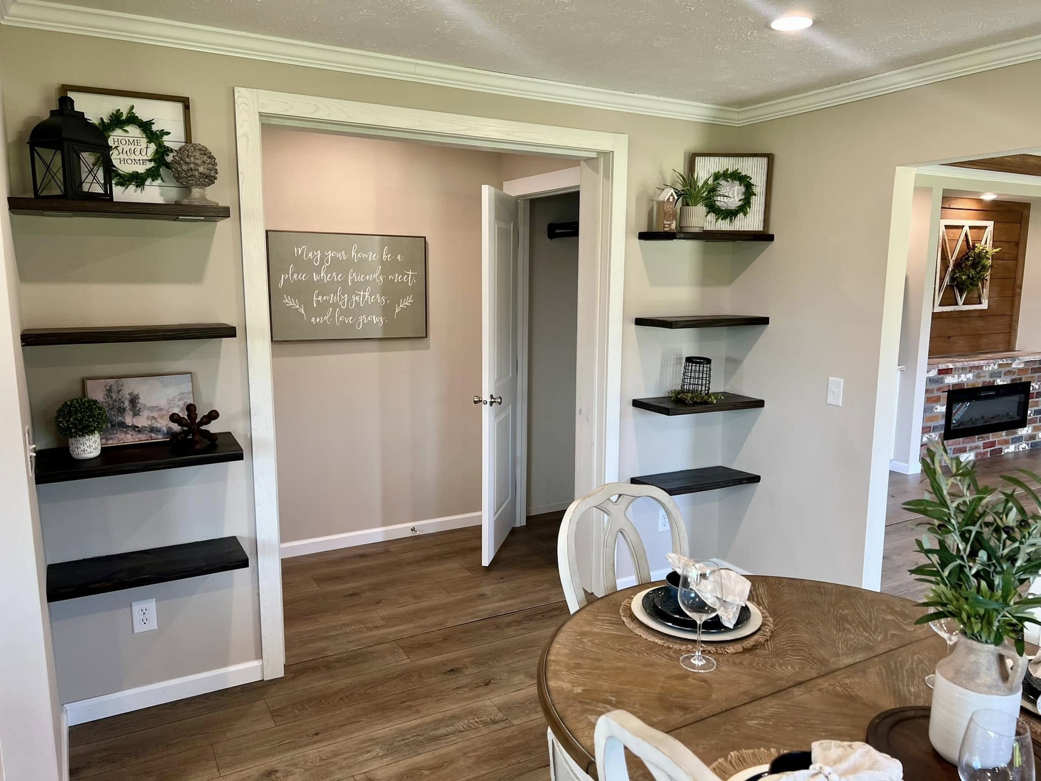 Cozy dining room with wooden table set. Shelves with greenery and decor frame an open doorway. Soft lighting and earthy tones create a warm, inviting atmosphere.