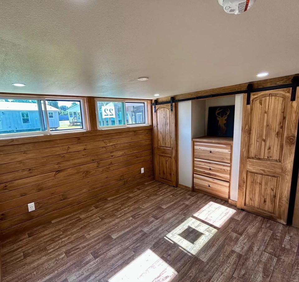 Cozy room with wood paneling, barn-style closet doors, and bright windows. A dresser and deer art add rustic charm. Sunlight casts a warm glow.