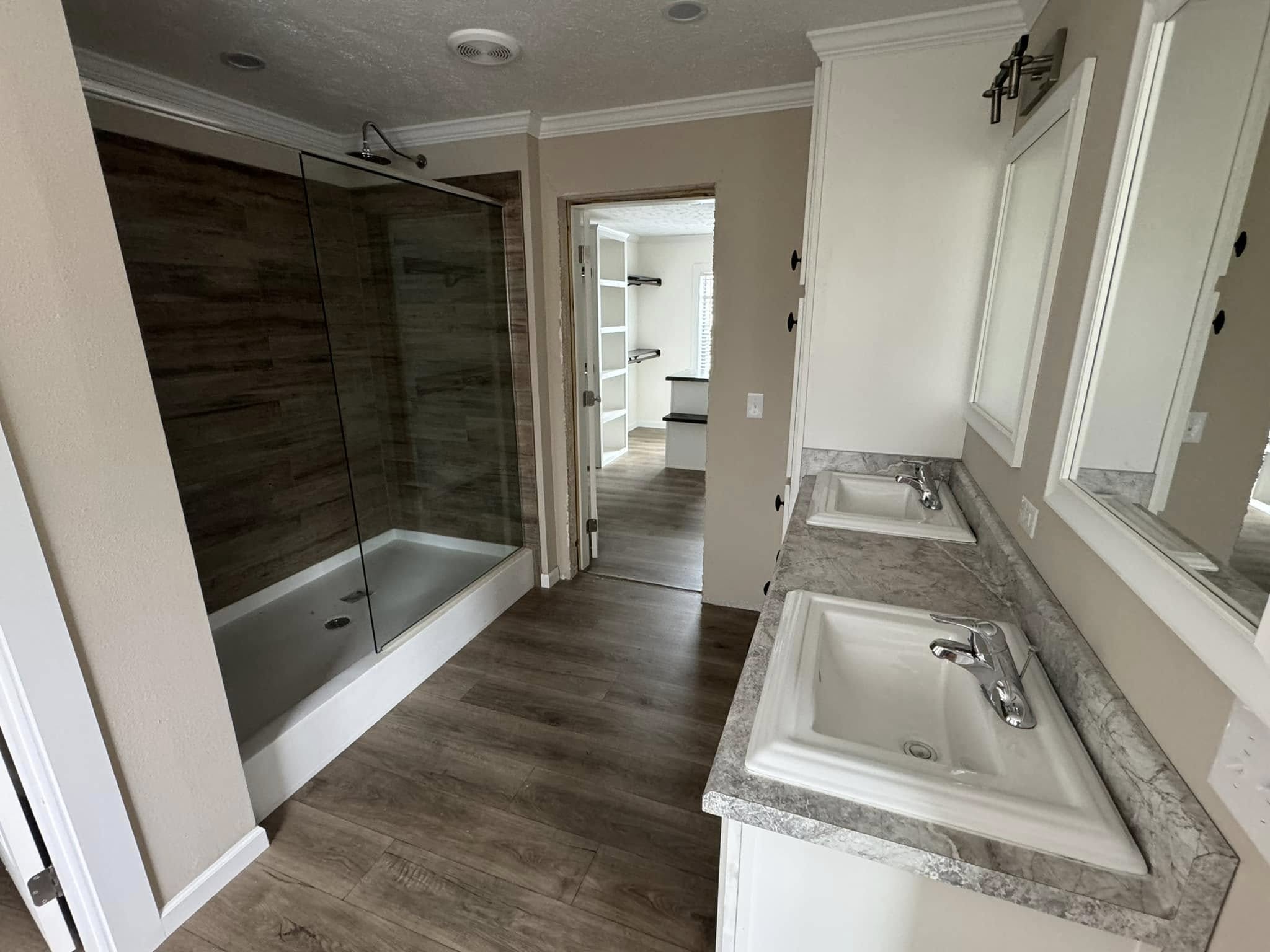 Spacious bathroom with light wood flooring, dual sinks on a marble countertop, and a glass-enclosed shower. Neutral tones create a calm, clean atmosphere.