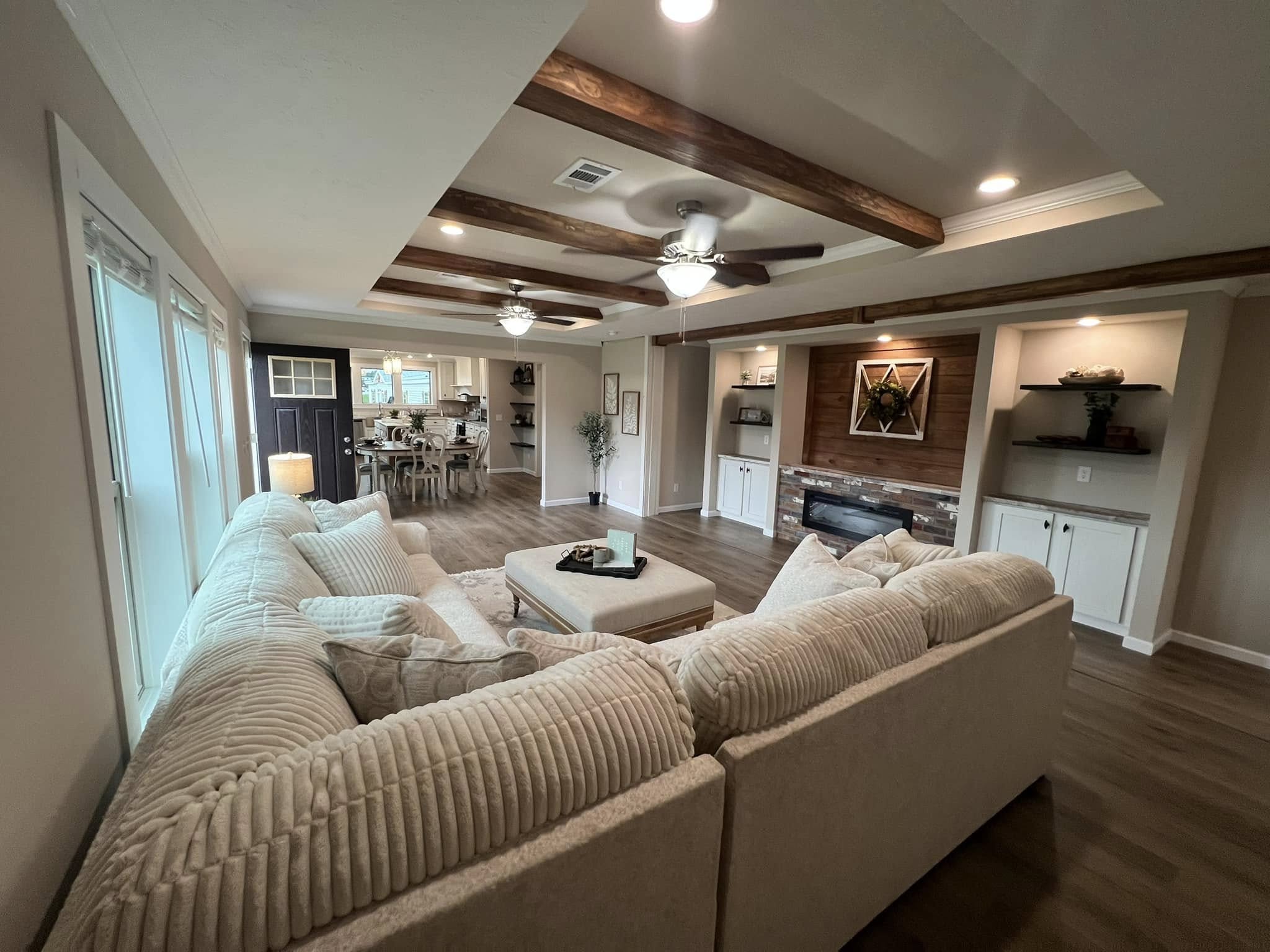 A cozy living room with a large beige sectional sofa facing a brick fireplace. Wooden beams and recessed lighting adorn the ceiling, while a dining area is visible in the background.