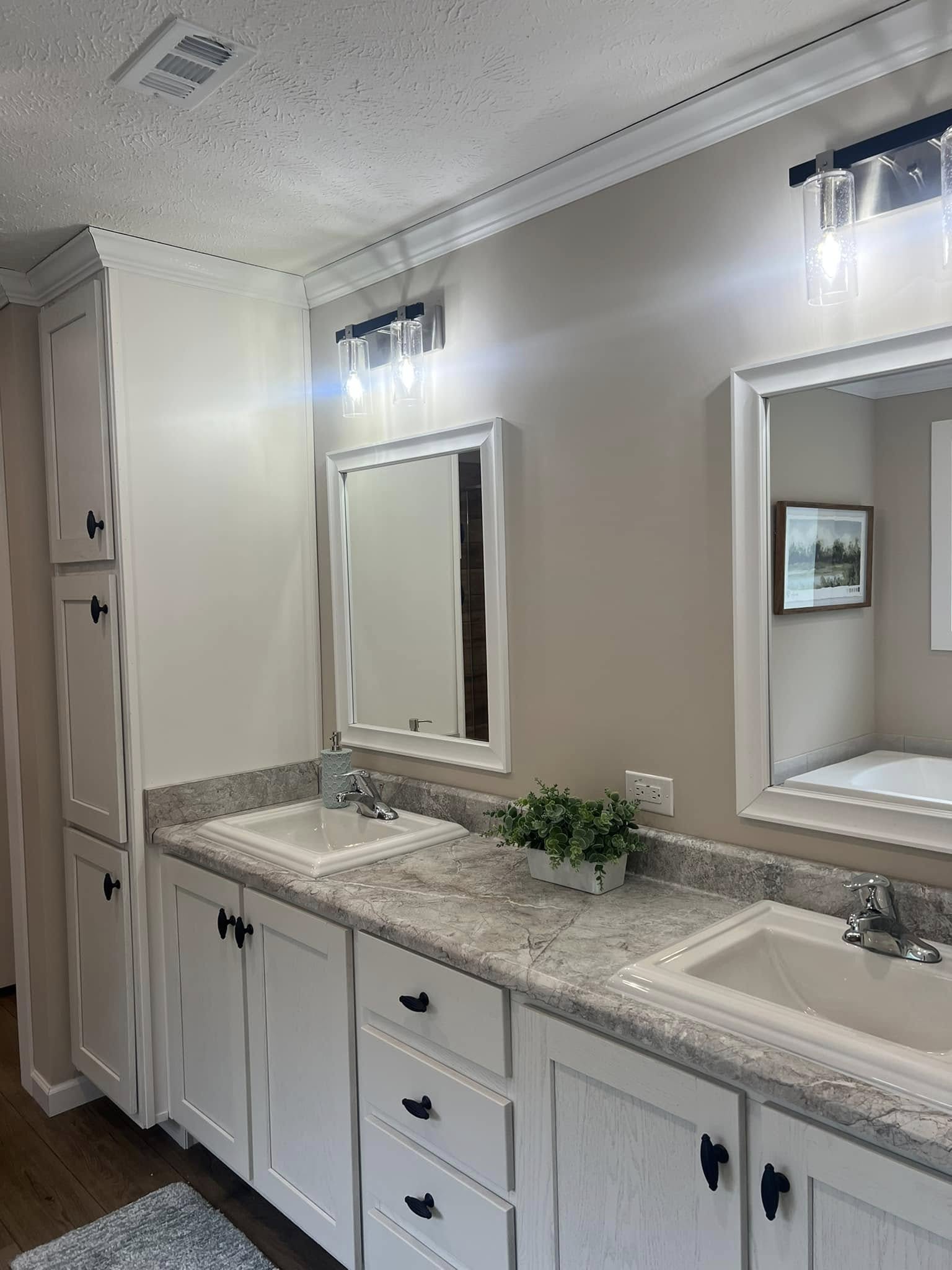 Bright bathroom with two sinks, light gray marble countertop, white cabinets, and rectangular mirrors. Modern light fixtures emit a cozy glow. A small potted plant adds greenery.