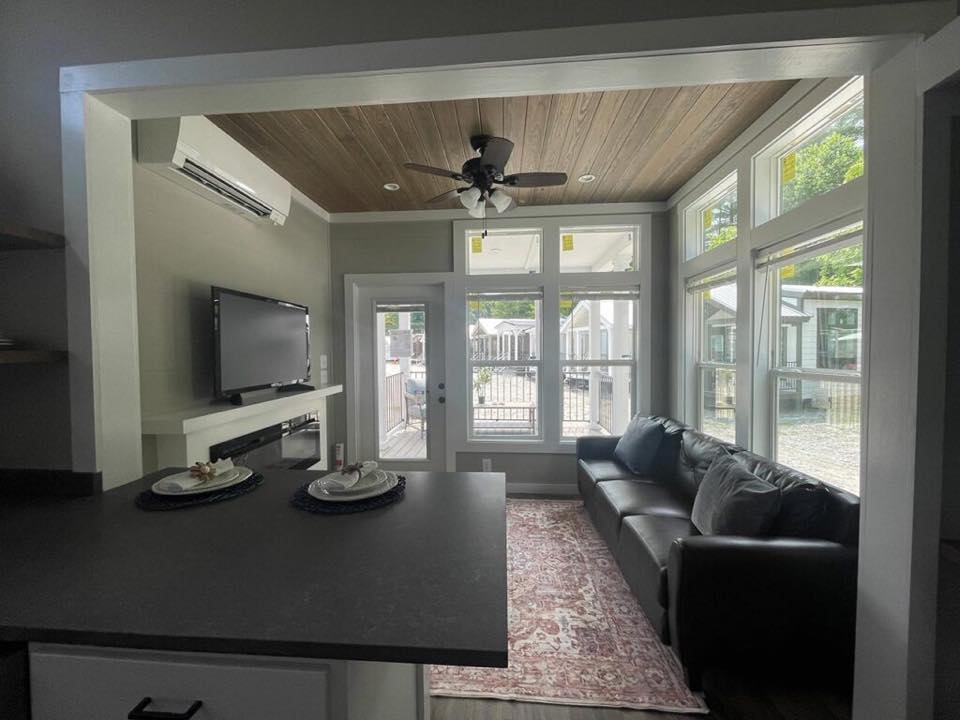 Cozy living room with dark sofa, wall-mounted TV, and ceiling fan. Large windows fill the room with natural light, creating an inviting atmosphere.