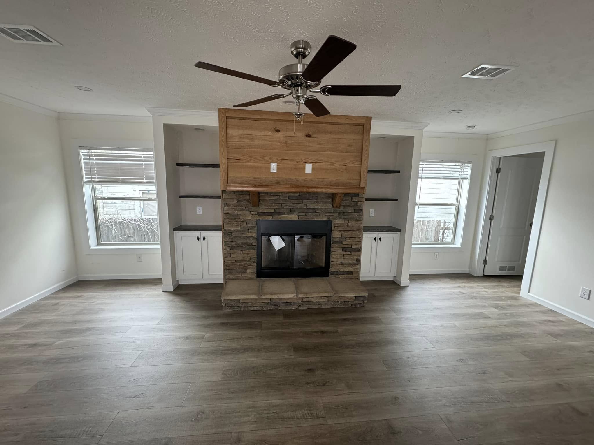 Spacious living room with wooden floors, stone fireplace, and ceiling fan. White walls, built-in shelves, and cabinets create a bright, inviting atmosphere.