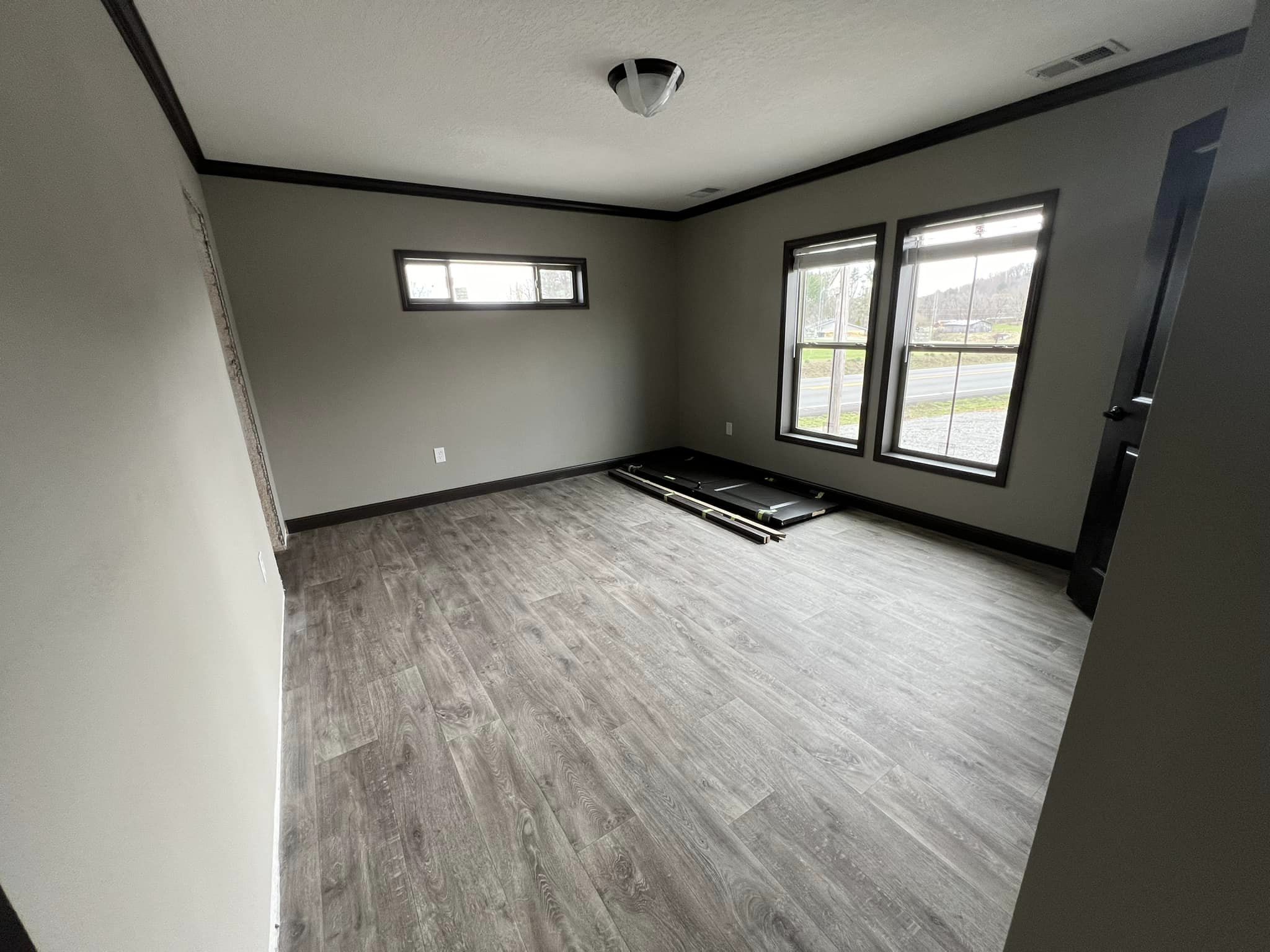 Empty room with light gray walls and wooden floor, featuring two tall windows and one narrow window. Bright ambiance with a stack of frames on the floor.