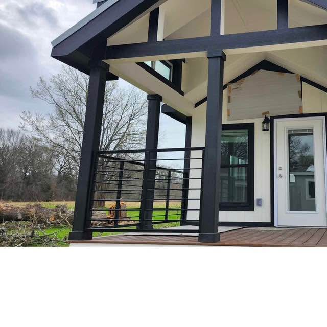 Modern tiny house with black trim and railing, featuring a wooden deck. Glass door and windows reflect a cloudy sky, surrounded by barren trees.