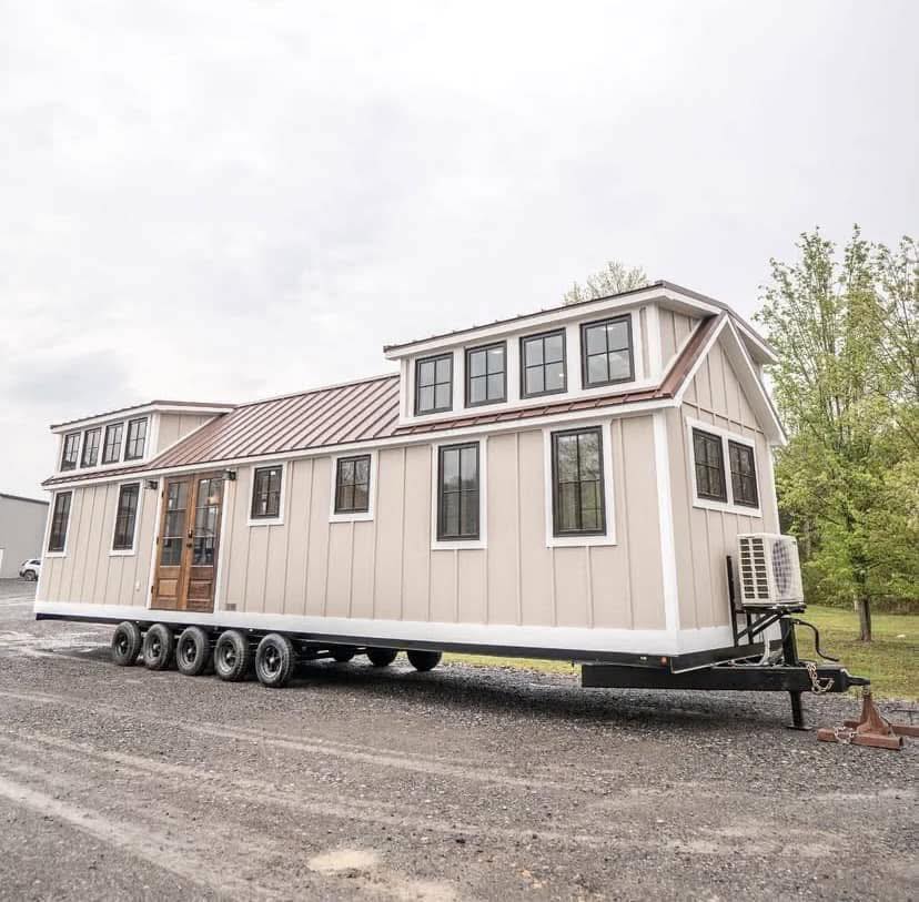 A beige tiny house on wheels with a sloped metal roof and multiple windows. It sits on a six-wheeled trailer, parked on a gravel surface near trees.