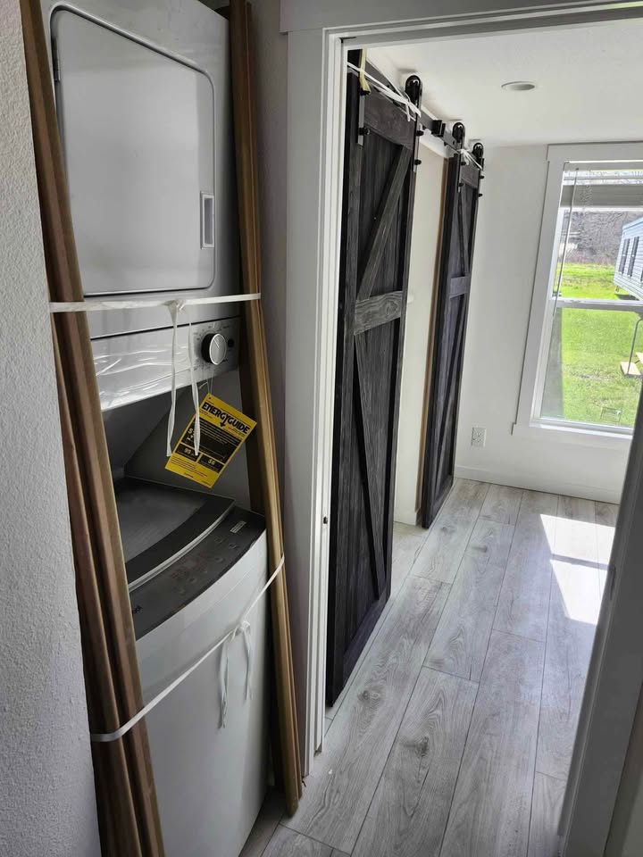 A narrow hallway features a white stacked washer and dryer unit on the left. It leads to rustic dark barn-style sliding doors, opening to a sunlit room with a large window. The scene feels cozy and modern.