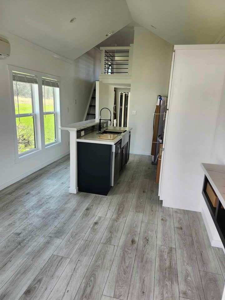 Modern tiny house interior featuring light wood flooring, a sleek black and white kitchen with an island, and a loft area accessed by stairs, creating an open, airy atmosphere.