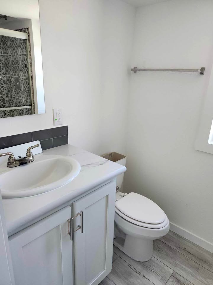 A small, modern bathroom with a white sink and cabinet, chrome faucet, toilet, and towel bar. Light gray tile floor and partial tiled mirror wall visible.