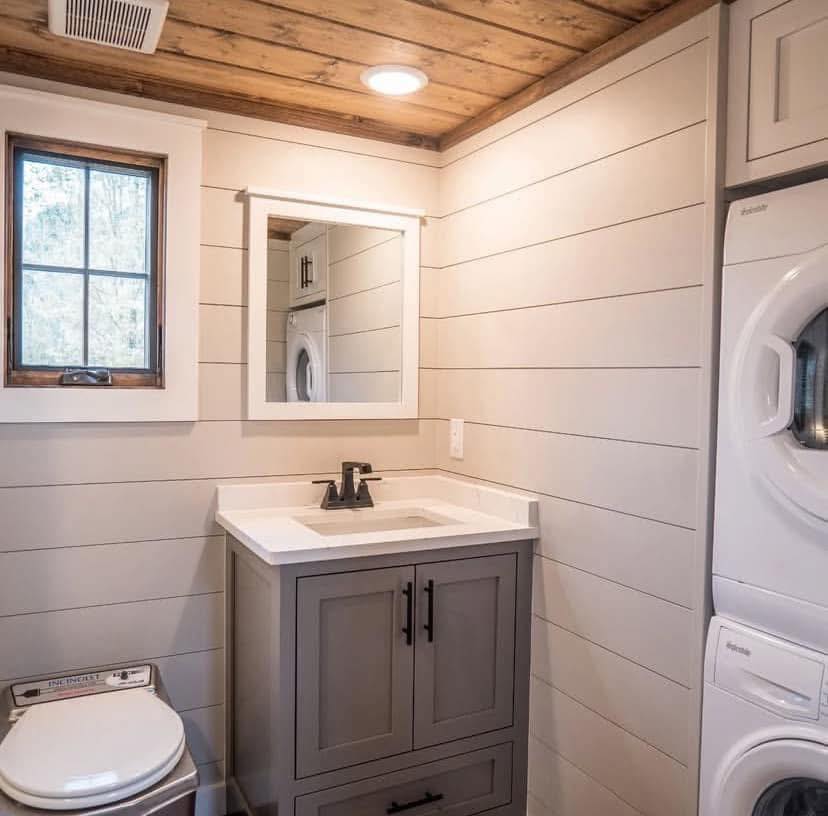 A small bathroom features a vanity with a rectangular mirror, shiplap walls, and a stackable washer and dryer. Wood ceiling adds warmth. Cozy and functional.