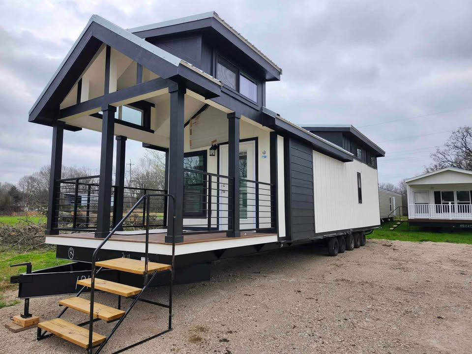 Modern tiny home on wheels with a gabled roof and large porch, set on a gravel surface against a cloudy sky, conveying a sense of mobility.