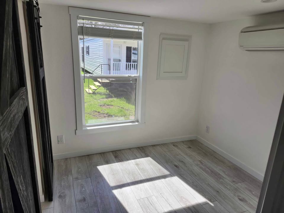 Bright, empty room with light gray wooden floors, a large window showing a green lawn and steps, a wall-mounted air conditioner, and a rustic sliding barn door.