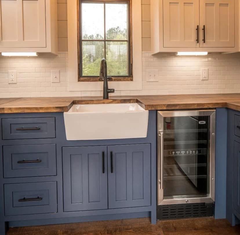A kitchen features blue cabinets with black handles, a white farmhouse sink, a wooden countertop, wine cooler, and a window with a view of greenery.