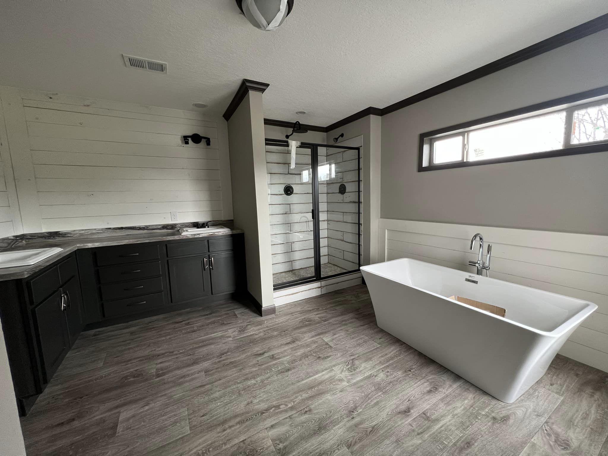 Spacious bathroom with rustic wood flooring, black cabinetry, glass shower, and a white freestanding tub under a rectangular window, conveying relaxation.