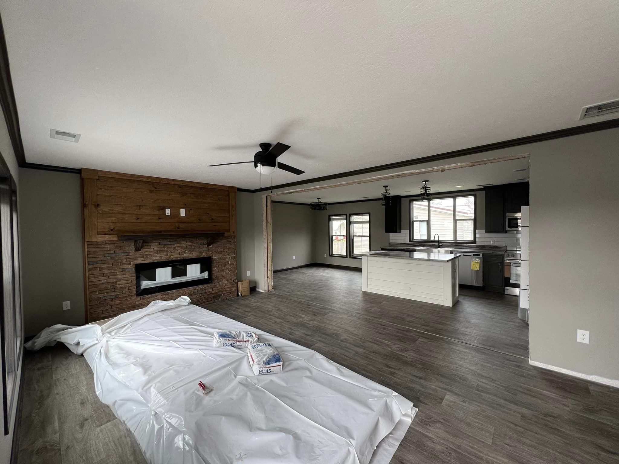 Open-concept living area with wood flooring, a stone fireplace with a wooden mantel, ceiling fans, and a modern kitchen with an island.