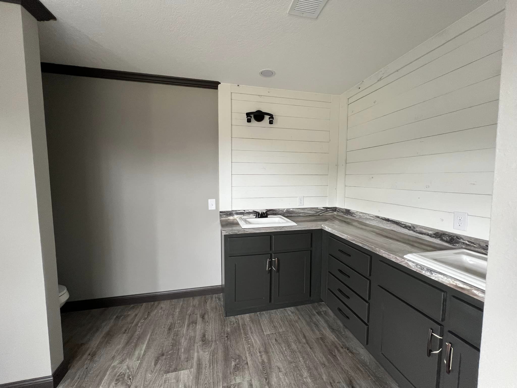 Small modern kitchenette with dark cabinets, gray countertops, and a shiplap wall. It features a sink and dark wood flooring, creating a minimalist feel.