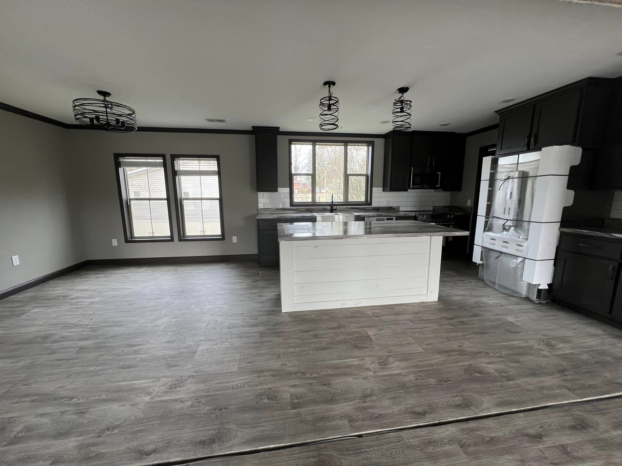 Spacious modern kitchen with dark cabinets, light grey island, and wooden flooring. Pendant lights hang above. Large windows enhance natural light.