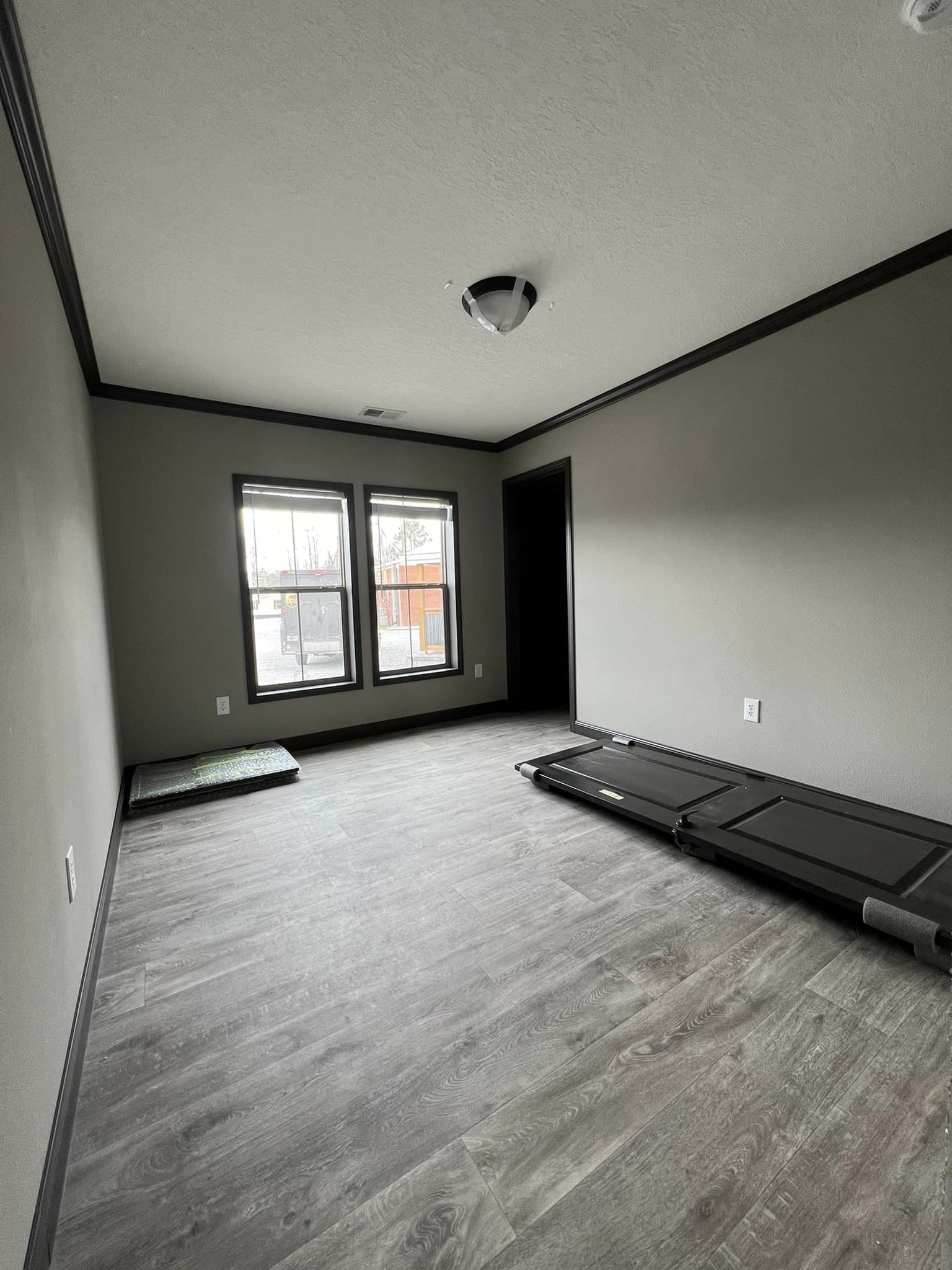A small, empty room with light gray walls and dark floor trim, featuring wooden flooring. A folded treadmill lies on the floor near two windows, allowing soft natural light to enter.
