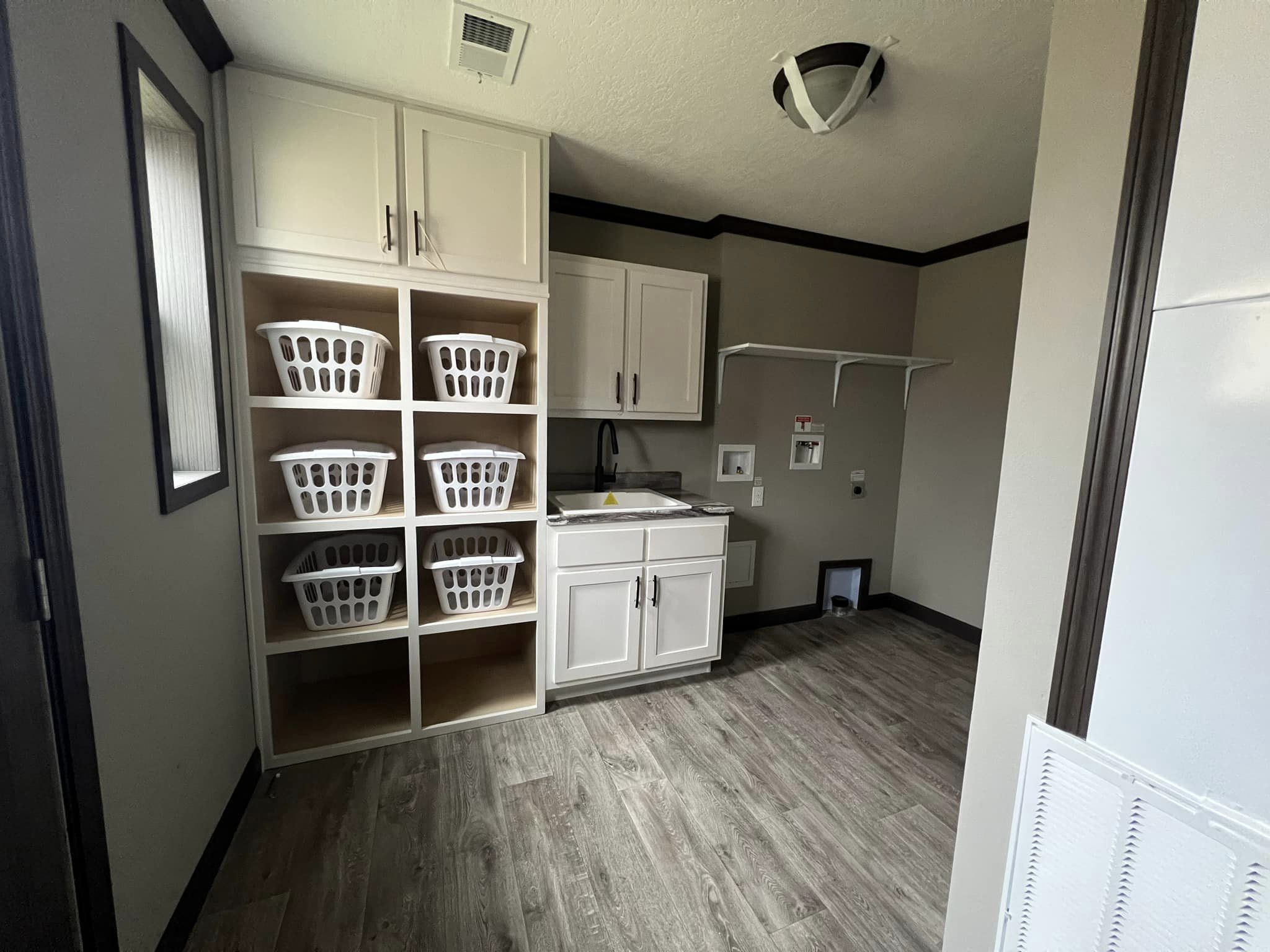 Spacious laundry room with six white laundry baskets on wooden shelves, a sink with white cabinets, laminate flooring, and neutral-colored walls.