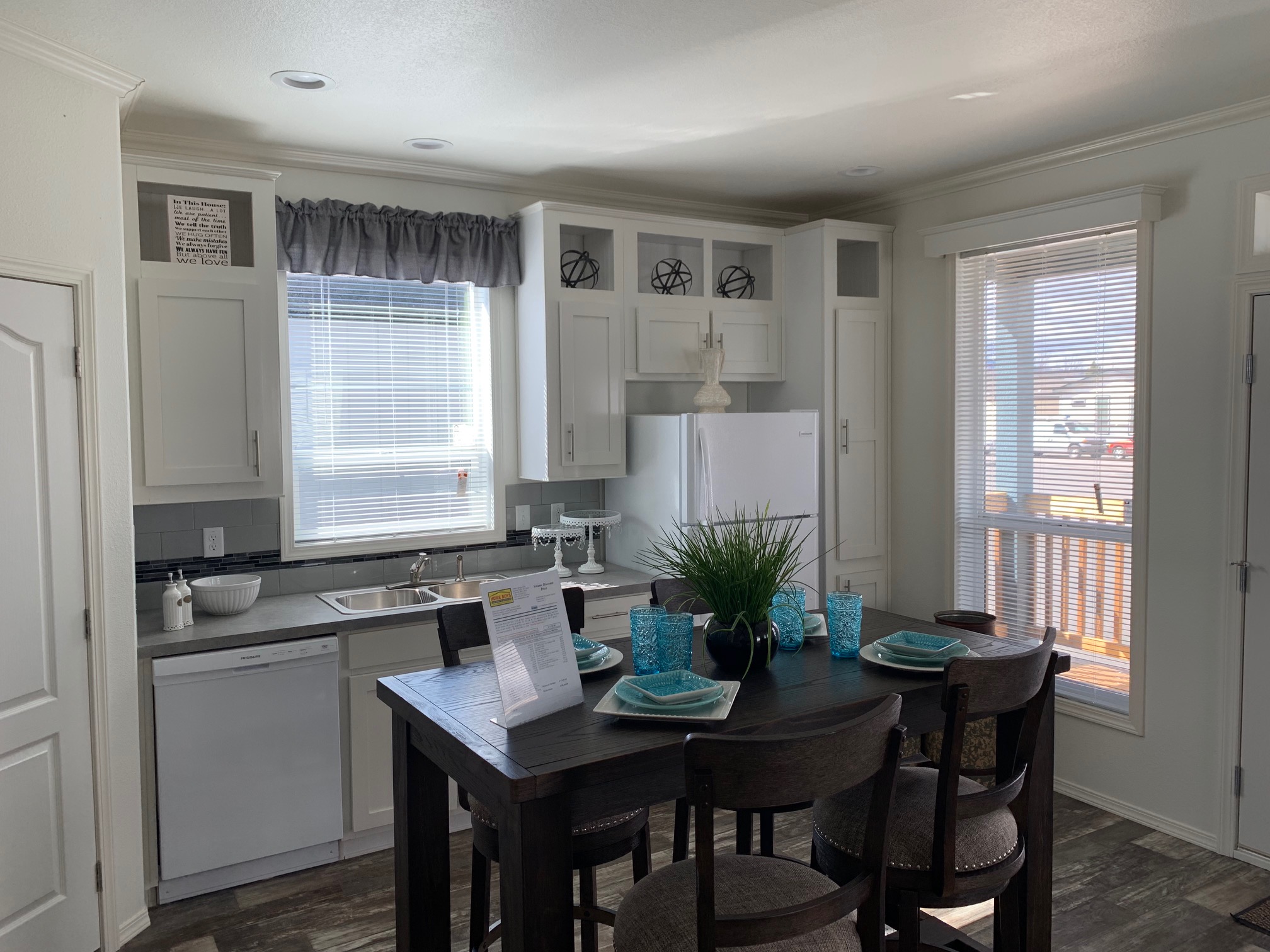 Bright kitchen with white cabinets and a large window, featuring a dark wood dining table set with turquoise plates and glasses. Cozy and inviting.