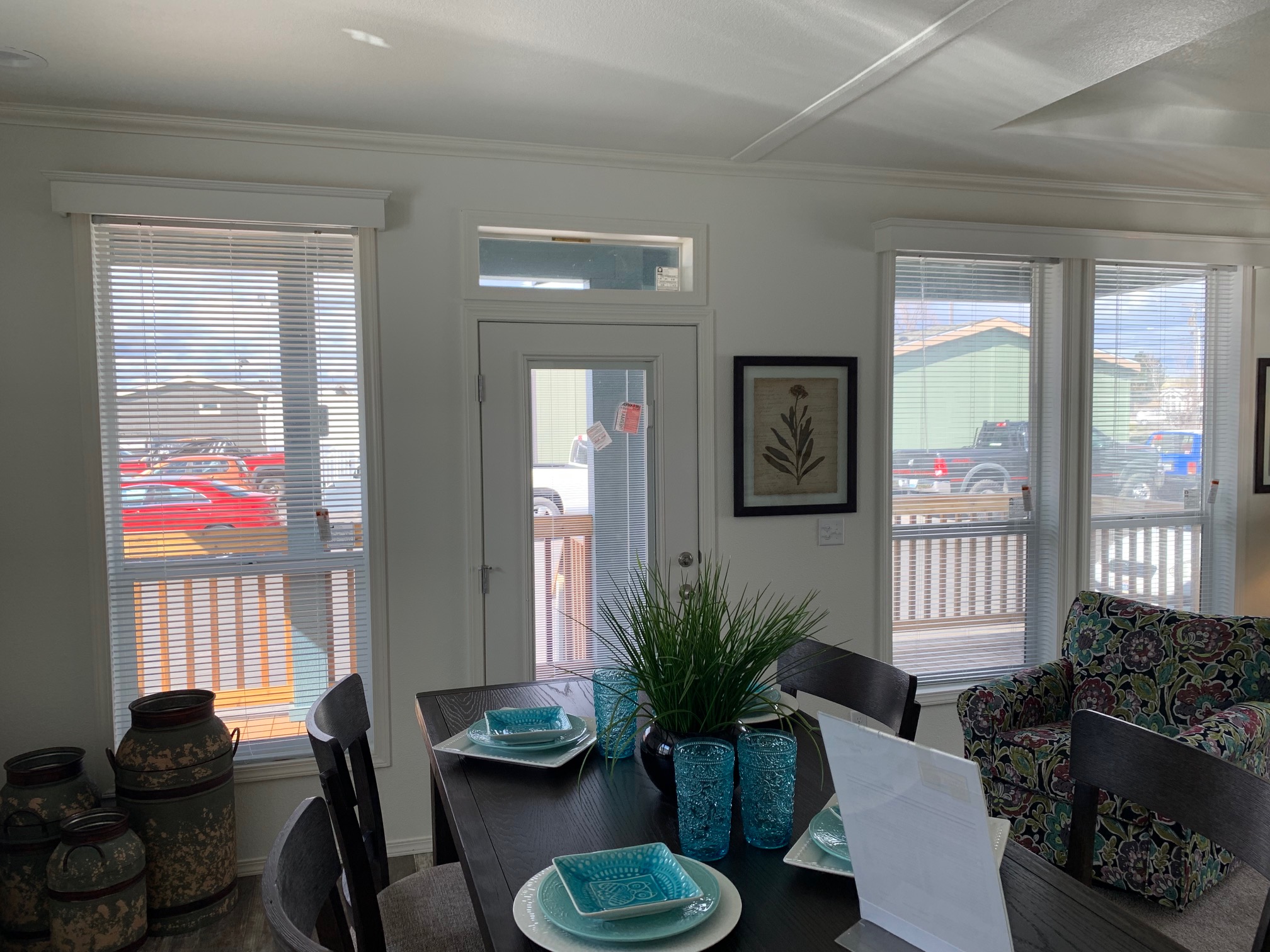 A bright dining area with a wooden table set for four, featuring teal plates and glasses. A plant centerpiece adds freshness. A floral-patterned chair is on the right, and sunlight streams through large windows, revealing cars outside, creating a cozy yet airy atmosphere.