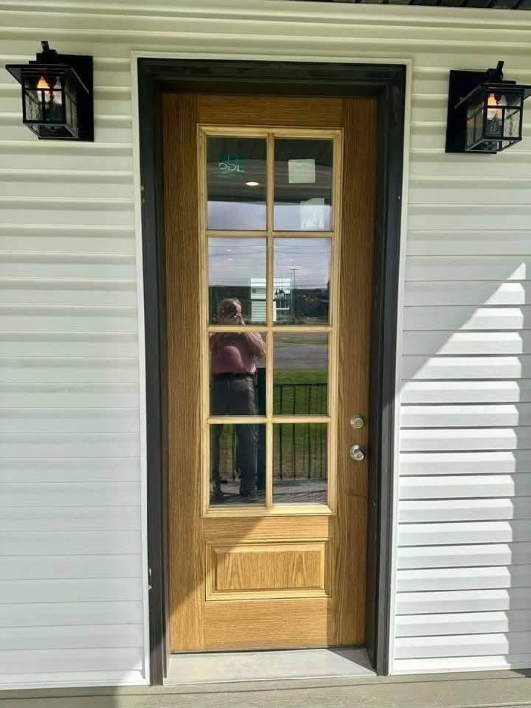 A glass-paneled wooden door with a reflection of a person in a pink shirt. Flanked by two black wall lanterns, set against white siding under bright sunlight.