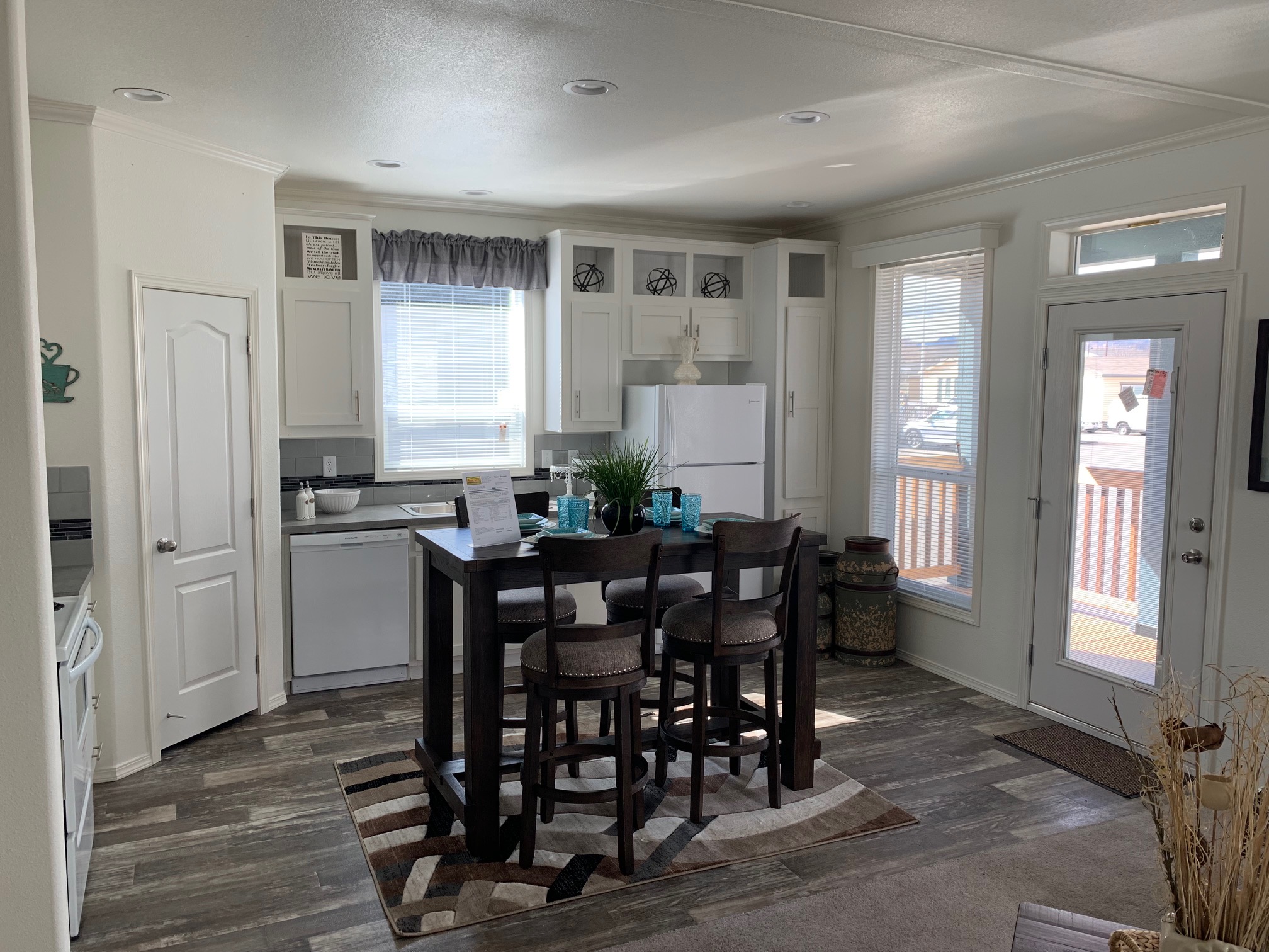 Bright kitchen with white cabinets, gray countertops, and wood floor. A dark wood table with four stools sits in the center. Light streams through the windows, creating a welcoming atmosphere.