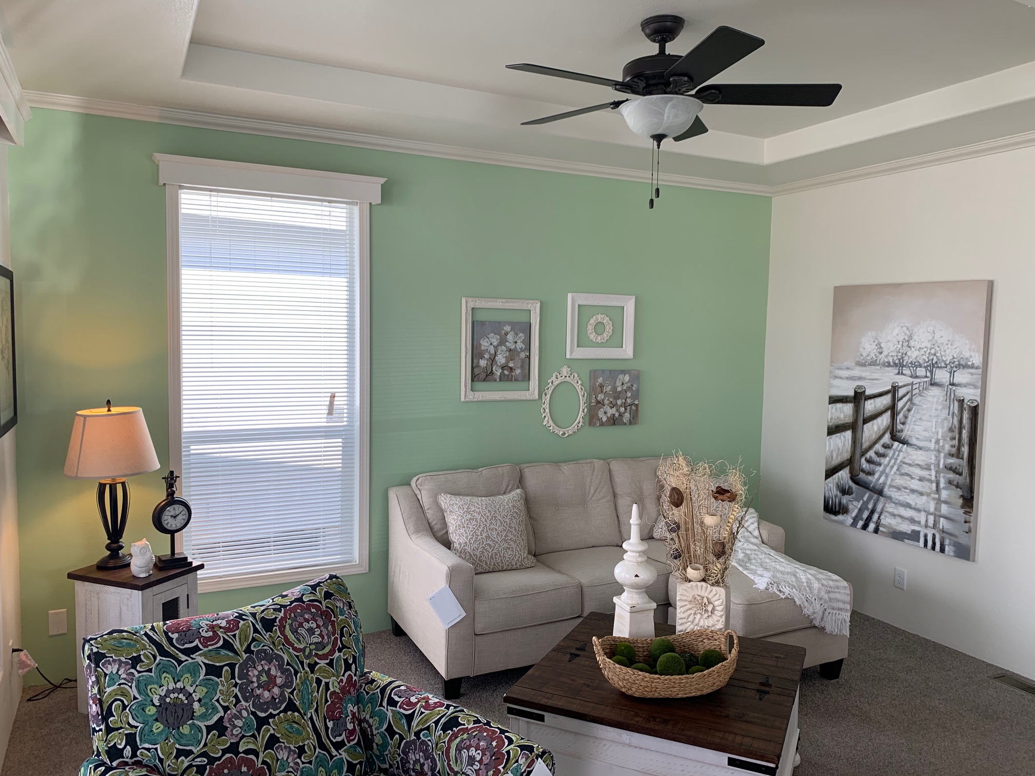 Cozy living room with a pastel green accent wall, cream sofa, floral armchair, and wood coffee table. Art and decor add a serene, welcoming feel.