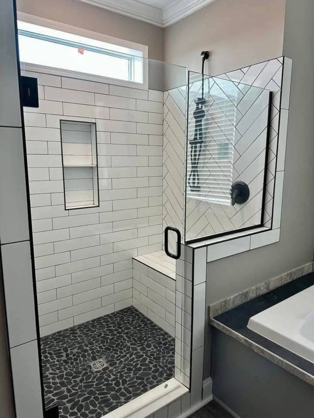 Modern bathroom shower with geometric white tile walls and stone floor. Glass enclosure with black trim and handle. Bright and clean ambiance.