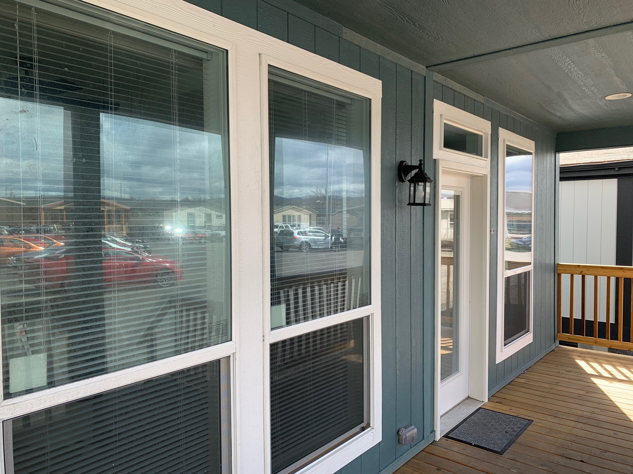 A porch with wooden flooring and blue siding has several large windows with white frames. A black outdoor light is mounted beside a white door.