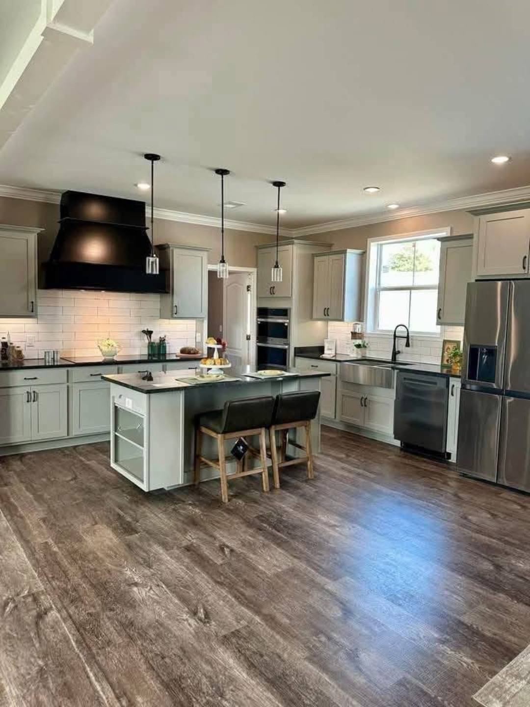 Modern kitchen with light gray cabinets, black range hood, and stainless steel appliances. Central island features two bar stools, under warm lighting.