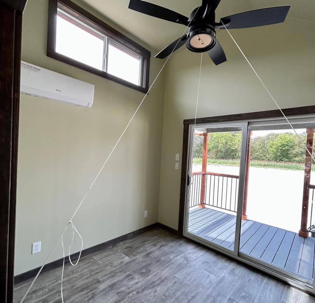 A small room with light green walls and dark wood trim features a black ceiling fan. There's a wall-mounted air conditioner and sliding glass doors leading to a patio with a view of greenery. Sunlight fills the space, creating a tranquil ambiance. A white string hangs from the ceiling to the floor.