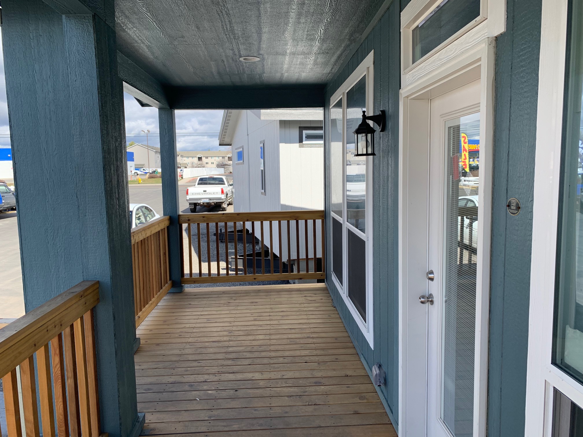 Covered porch with wooden decking and blue walls. A white door with glass panes is on the right. The railing overlooks a parking lot. Casual and inviting.