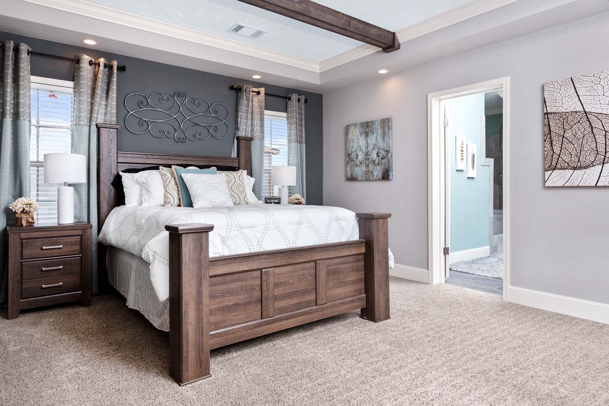 Cozy bedroom with a dark wood bed, patterned pillows, and light carpet. Gray walls, decorative art, and soft lighting create a calm atmosphere.