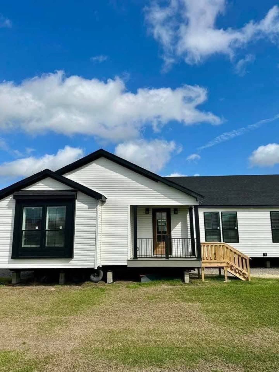Single-story, white modular home with black trim, elevated on concrete blocks. Front porch with a wooden ADA ramp. Bright blue sky, scattered clouds above.