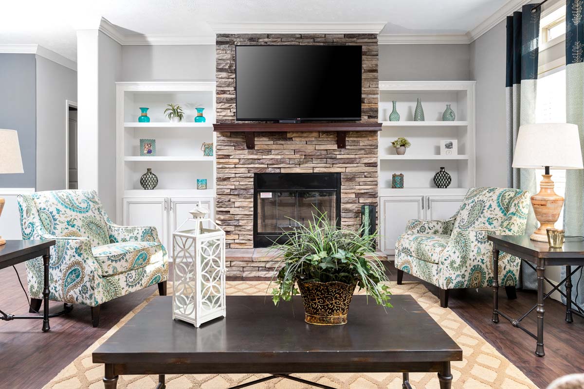 Elegant living room with a stone fireplace, flanked by white shelves holding teal decor. Two patterned chairs face a TV. A dark coffee table with a plant centers the space.