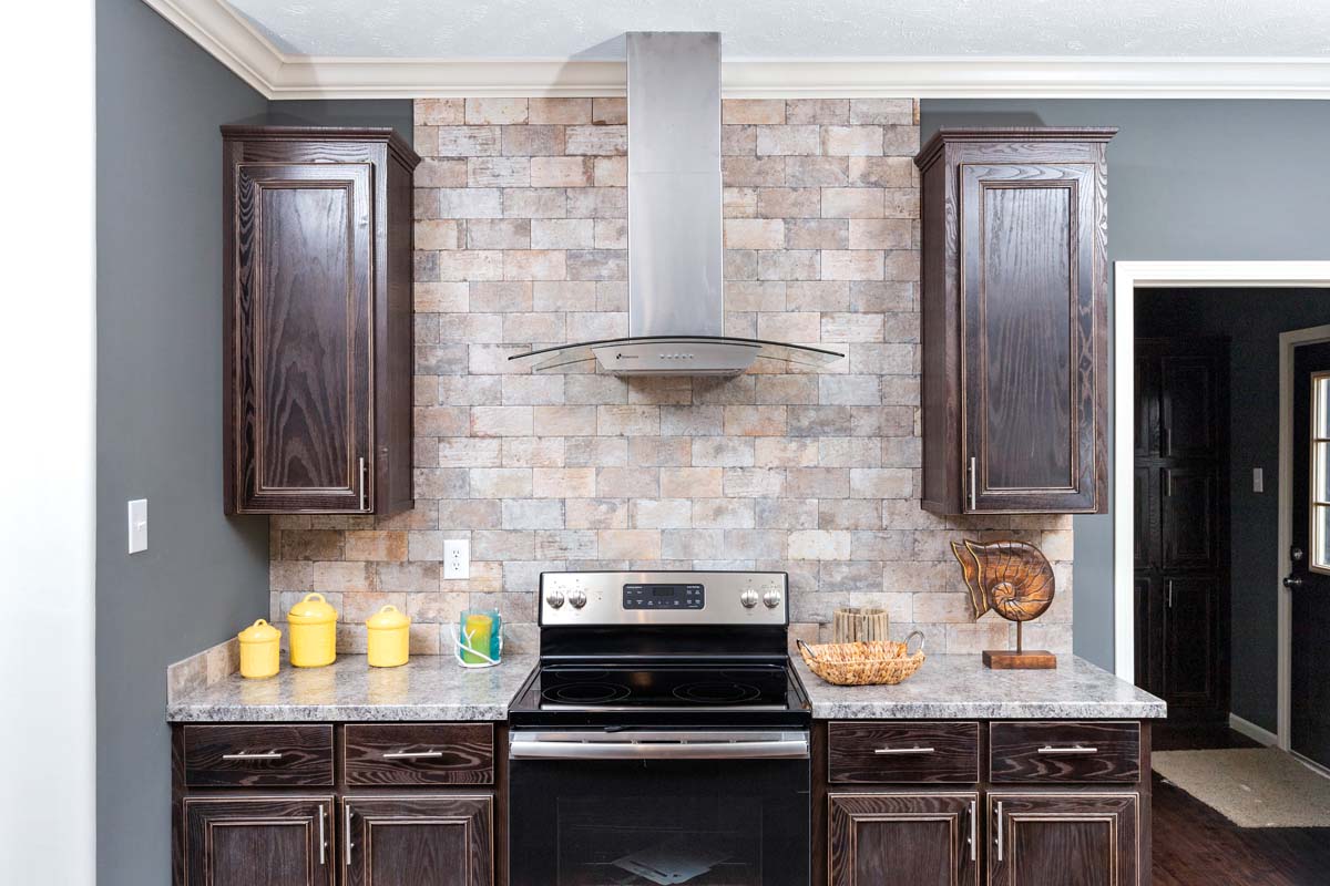 Modern kitchen with dark wood cabinets and a stainless steel stove. A stone tile backsplash adds a rustic touch. Yellow canisters and decorative items adorn the countertop.