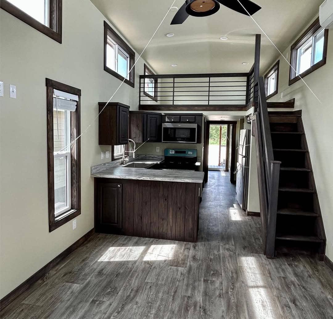 Open-concept tiny home interior with a modern kitchen, loft, dark wood cabinetry, and a staircase. Natural light from multiple windows enhances the airy feel.