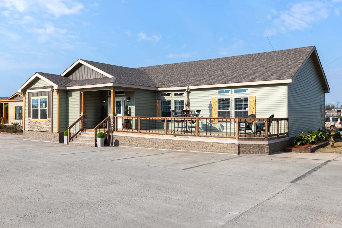 A modern, single-story house featuring pale green siding, a stone-accented facade, and a spacious front deck. Clear skies enhance the inviting, tranquil setting.